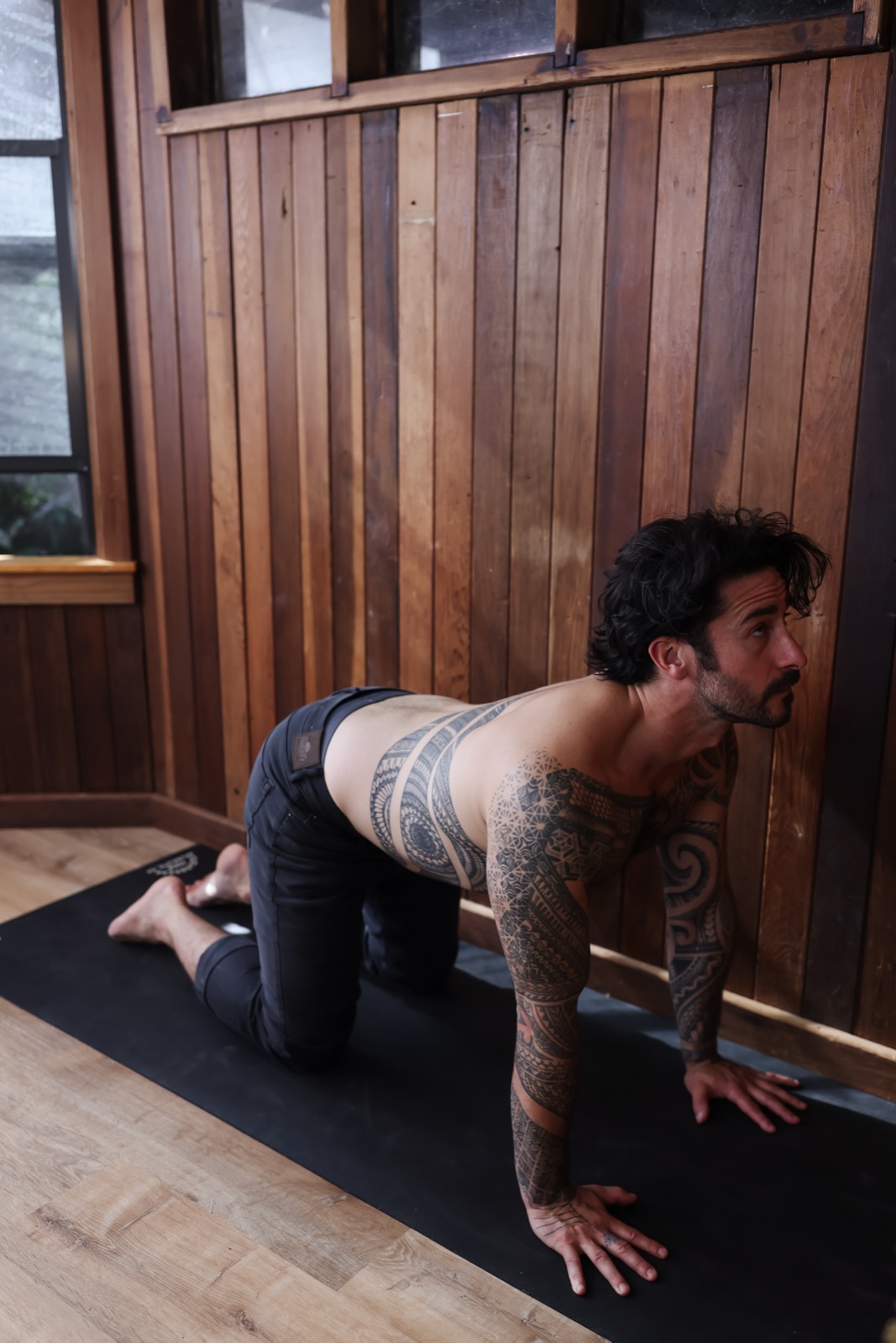 Peter shirtless in cat pose on a mat against wood paneling, tattoos visible.
