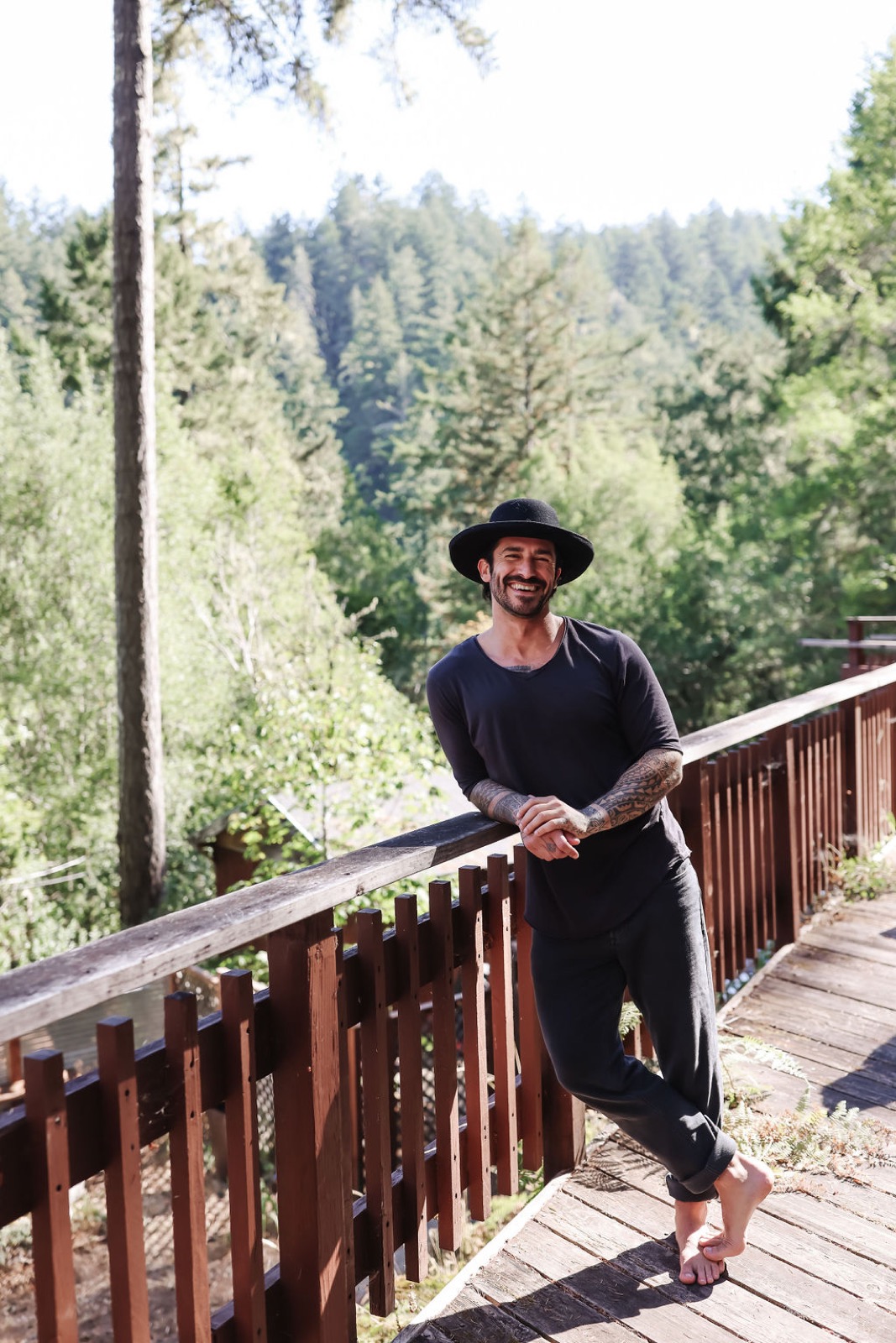 Peter in a wide-brim hat leaning on a forest deck railing, smiling.